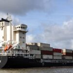 Container ship navigating Hamburg harbor with stacked cargo under a cloudy sky.
