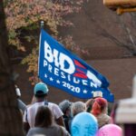 A diverse crowd gathered outdoors holding a Biden 2020 flag during a protest.