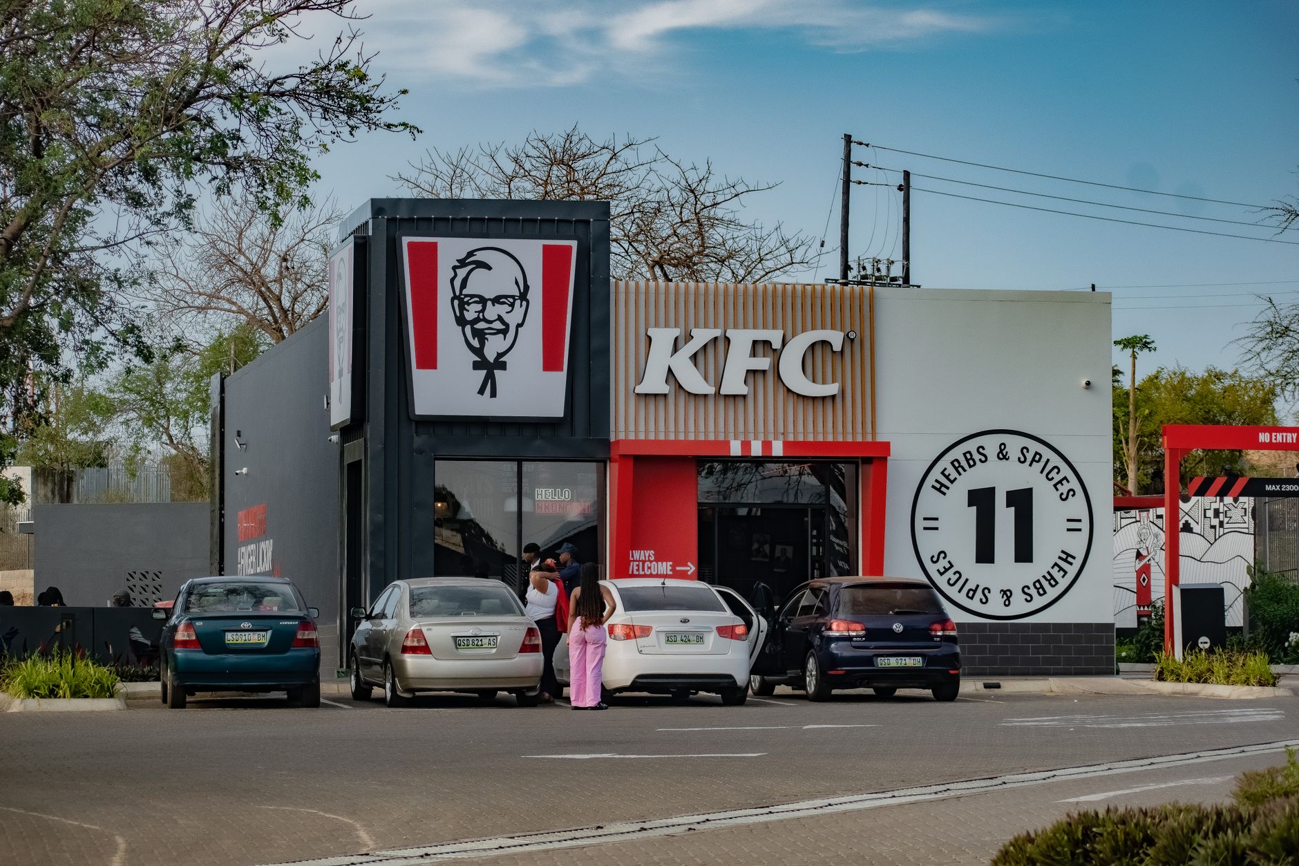 Street view of a KFC restaurant in Manzini, Eswatini, showing parked cars and patrons entering.