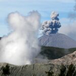 A dramatic aerial view of an erupting volcano with thick smoke plumes and rugged landscape.