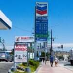 People walking near a Chevron gas station with prominent signage and parked vehicles on a sunny day.