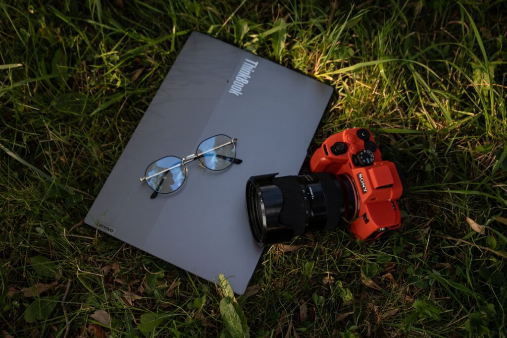 A laptop, camera, and glasses rest on grass, blending technology and nature.