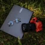 A laptop, camera, and glasses rest on grass, blending technology and nature.