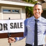 Smiling real estate agent with a for sale sign in front of a house.
