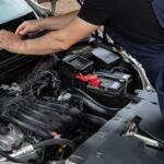 Mechanic performing maintenance on a car engine outdoors during the day.