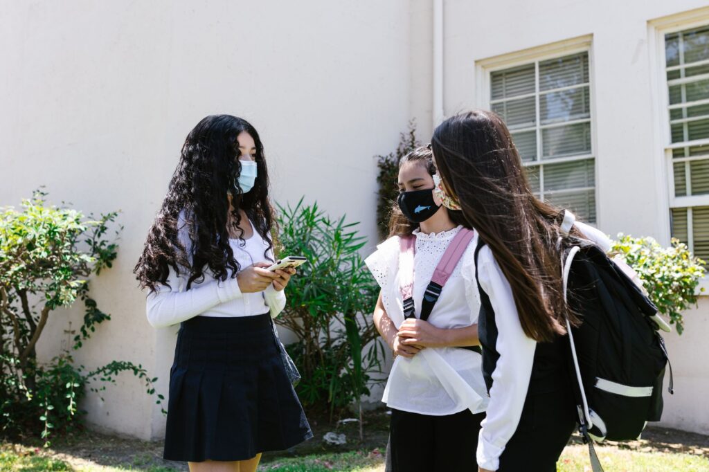 Teenage girls in face masks talking outside a school building, carrying backpacks and interacting outdoors.