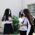 Teenage girls in face masks talking outside a school building, carrying backpacks and interacting outdoors.