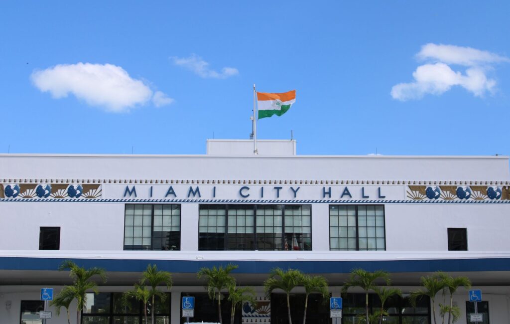 A clear view of Miami City Hall displaying an Indian flag under a bright blue sky.