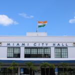 A clear view of Miami City Hall displaying an Indian flag under a bright blue sky.