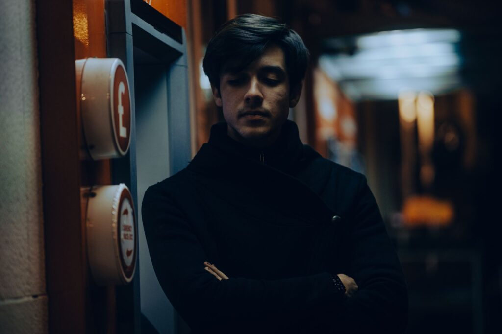 Thoughtful young man in a dark coat standing indoors at night.