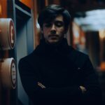 Thoughtful young man in a dark coat standing indoors at night.