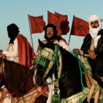 Traditional Moroccan horsemen in rich attire against a backdrop of Moroccan flags at dusk.