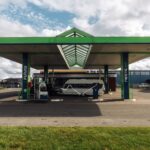 A modern gas station with a green canopy and parked camper van under a cloudy sky.