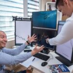 Two businessmen enthusiastically analyzing market charts on computer monitors in a modern office.