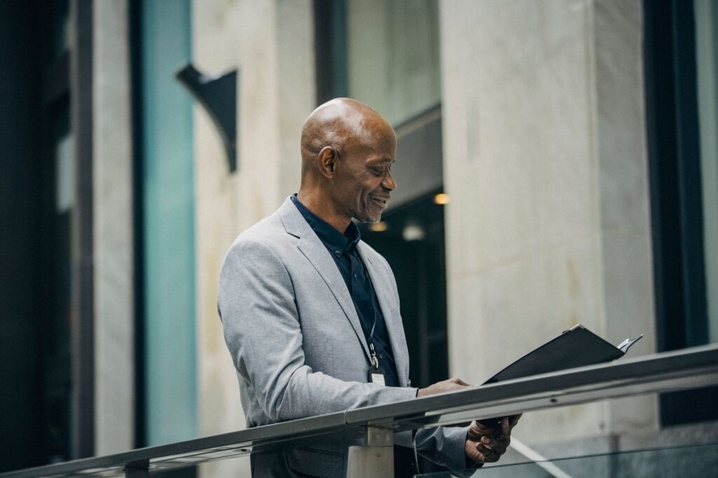 SIde view of middle aged smiling black executive in formal suit standing near fence and reading papers in card board near modern office