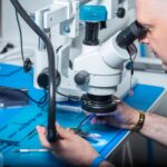 Technician examining electronic chip under a microscope in a computer lab setting.