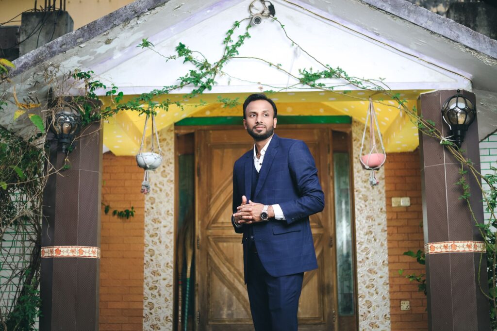 Man in a blue suit standing in front of a doorway, surrounded by greenery and hanging planters.