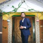 Man in a blue suit standing in front of a doorway, surrounded by greenery and hanging planters.