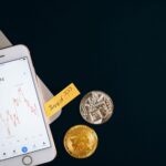 A smartphone displaying cryptocurrency prices beside coins and a laptop, symbolizing investment.