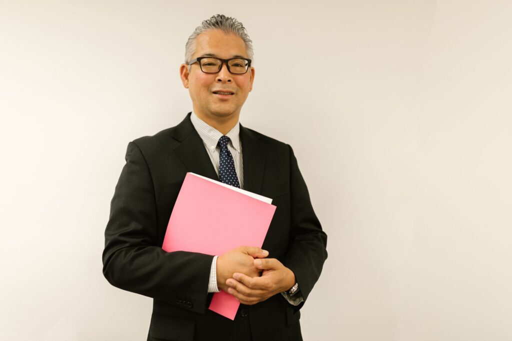 Confident businessman in black suit holding pink folder, smiling at the camera indoors.