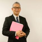Confident businessman in black suit holding pink folder, smiling at the camera indoors.