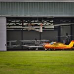 A vibrant yellow plane parked outside a hangar full of aircrafts on a sunny day.