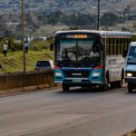 Street scene showing buses on a road with hilly landscape in the background.