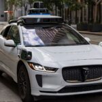 A white autonomous vehicle navigating a city street, reflecting urban architecture in daylight.
