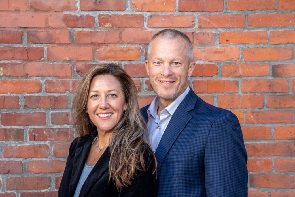 A cheerful middle-aged man and woman in business attire posing against a brick wall, depicting professionalism and partnership.