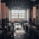 An empty cafeteria featuring wooden tables and chairs, lit by natural light through large windows.