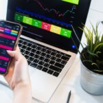 A person using a smartphone app to trade stocks with a laptop displaying market data.