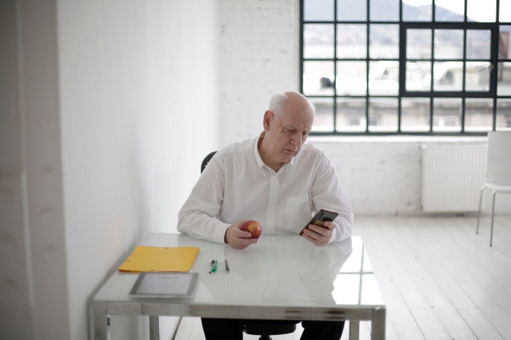 Elderly man in white shirt holding apple and messaging on mobile phone during short break in contemporary office