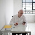 Elderly man in white shirt holding apple and messaging on mobile phone during short break in contemporary office