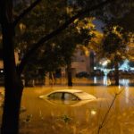 A dramatic scene of submerged cars in a flooded street surrounded by trees at night.