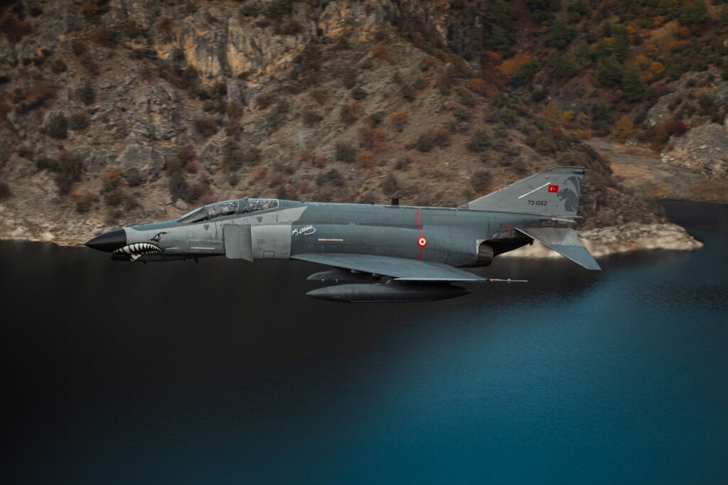 Turkish F-4 Phantom fighter jet flying over a scenic lake and cliffs in Türkiye.