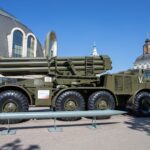 A military missile launcher on exhibit outdoors with a backdrop of unique architecture and clear blue sky.