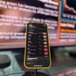 Smartphone showing stock market trends in front of blurred financial news screen indoors.
