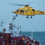 A yellow rescue helicopter performs a maritime operation over a cargo ship at sea.