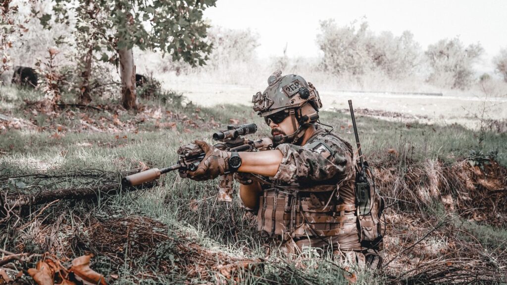 Military personnel in action, wearing camouflage and aiming a rifle outdoors.