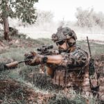 Military personnel in action, wearing camouflage and aiming a rifle outdoors.