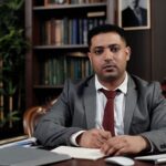 A serious businessman in a suit writing at his desk in a formal office setting.