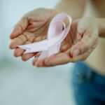 Crop unrecognizable topless lady demonstrating pink ribbon in arms as symbol of breast cancer awareness while standing in bright room
