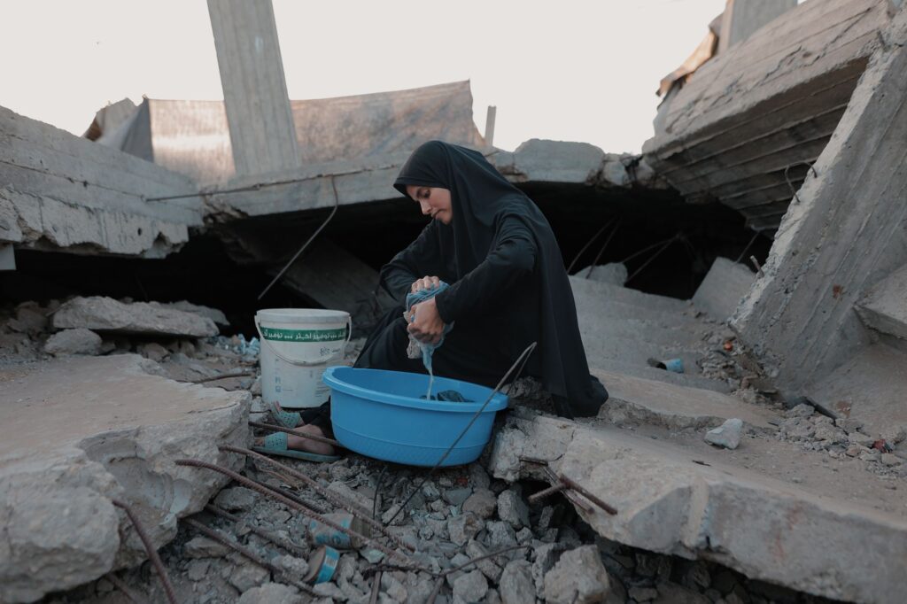 A woman in traditional attire washes clothes among the rubble in Gaza, depicting resilience.