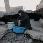A woman in traditional attire washes clothes among the rubble in Gaza, depicting resilience.