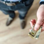 A close-up of a man's hand holding rolled US dollar bills, offering cash.