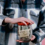 Close-up of a woman holding a savings jar labeled 'Where to next?' filled with US dollar bills.