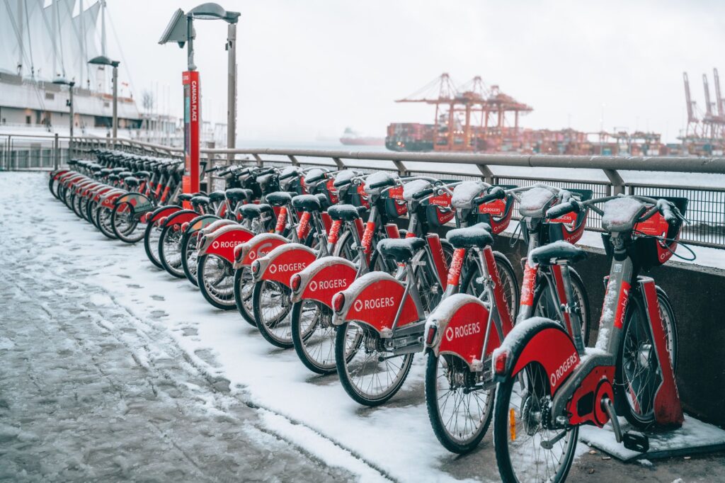 Snow-covered rental bicycles lined up by a waterfront with cranes in the background.
