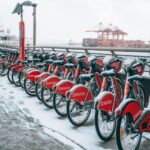 Snow-covered rental bicycles lined up by a waterfront with cranes in the background.