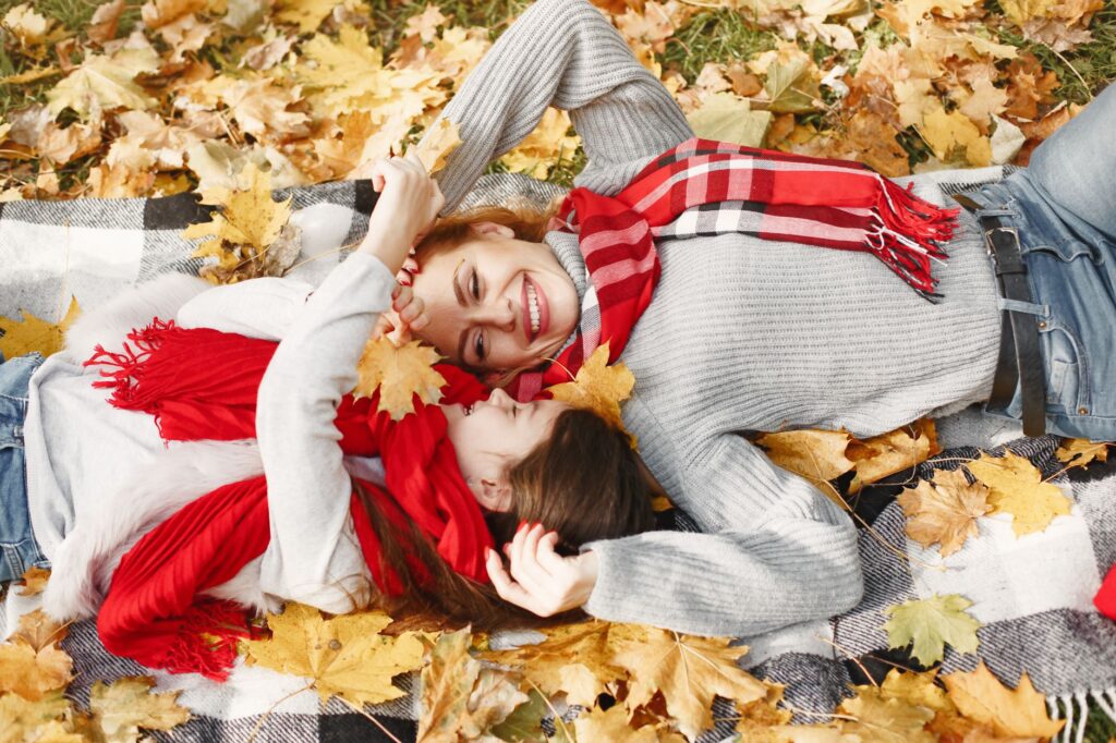 A joyful mother and daughter lying in autumn leaves, wearing red scarves and gray sweaters, smiling under the fall foliage.
