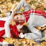 A joyful mother and daughter lying in autumn leaves, wearing red scarves and gray sweaters, smiling under the fall foliage.